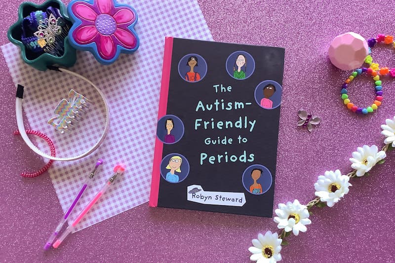 flat lay image of hardcover book 'the autism friendly guide to periods' on a pink background surrounded by hair accessories, pens, and daisies