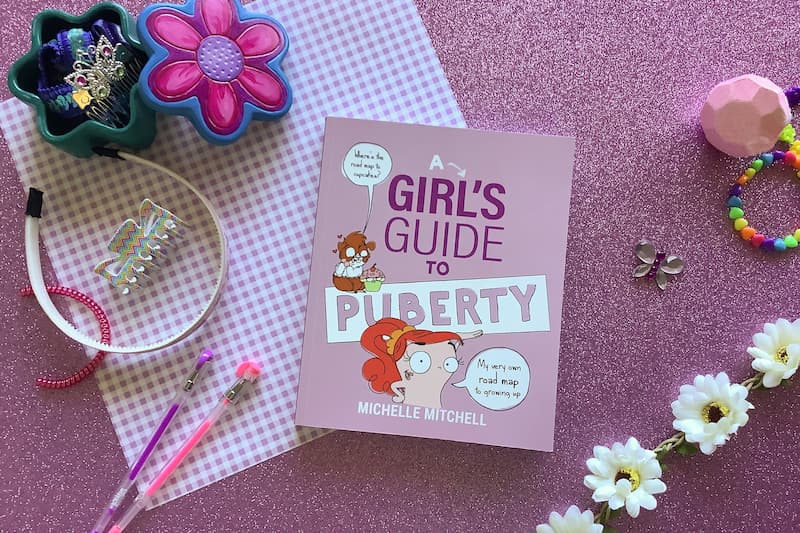 flat lay image of softcover book 'girls guide to puberty' on a pink background surrounded by hair accessories, pens, and daisies