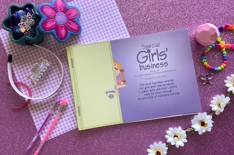 flat lay image of book 'special girls' business' on a pink background surrounded by hair accessories, pens, and daisies