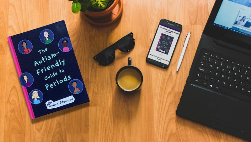 the book 'the autism-friendly guide to periods' sits on a desktop next to a pair of sunglasses and a cup of coffee. There is also a laptop and a mobile phone displaying the autism friendly periods web page.