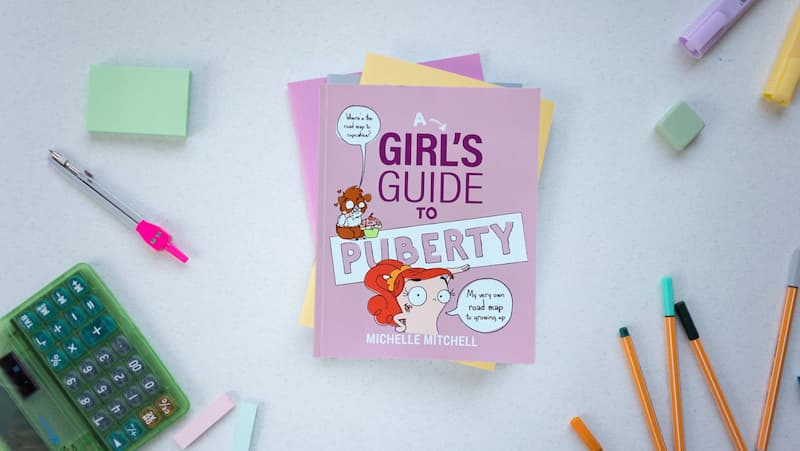 a stack of books sits on a desk surrounded by various stationery items. the top book is titled 'a girls guide to puberty'.